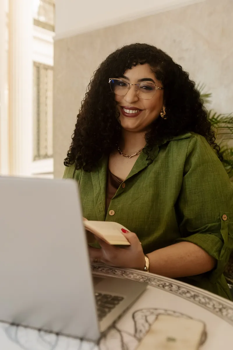 Camyla sorrindo para a câmera, com um livro nas mãos e o notebook à frente
